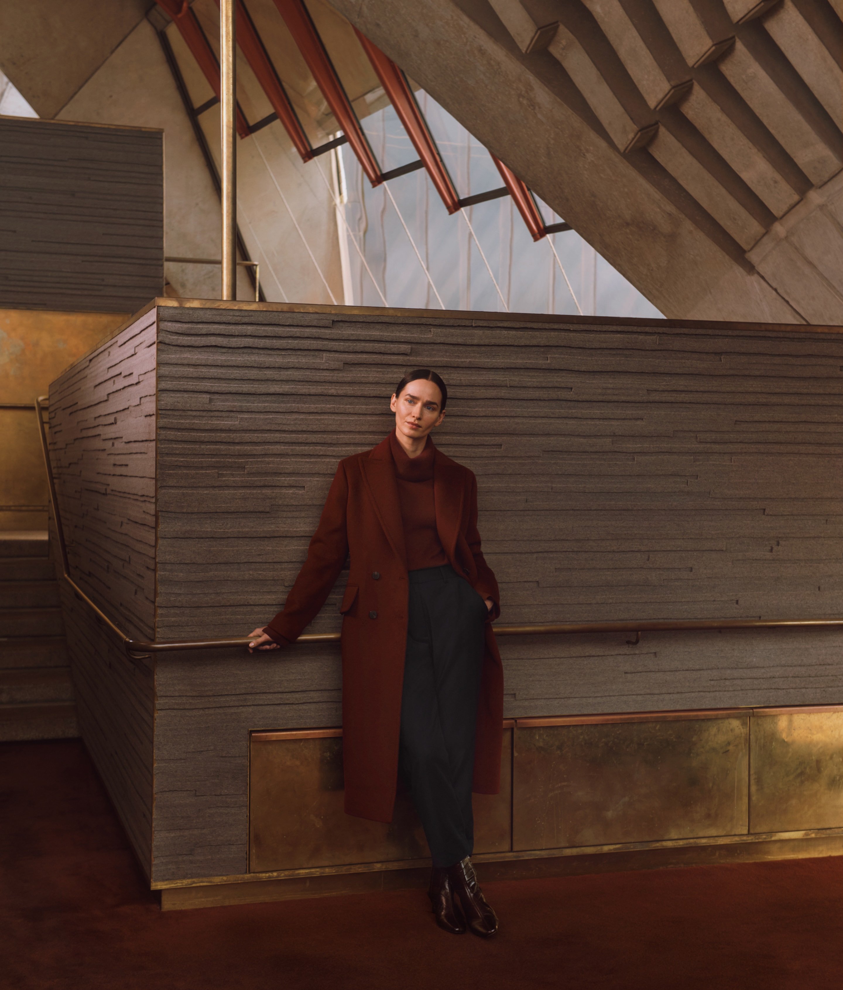 A fashionable adult female wearing a long-sleeved top, dark slacks, and a red overcoat, leaning against a concrete architectural wall indoors.
