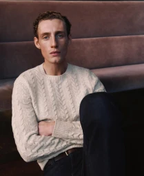 A man wearing a cream cable-knit sweater sits with arms crossed in front of a textured brown banquette.