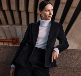 Woman wearing a dark corduroy blazer over a white turtleneck sweater leaning against a textured stone ledge.
