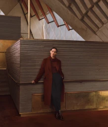 Model standing indoors wearing a burgundy coat and dark trousers against architectural walls.