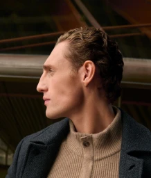 Profile portrait of a man wearing a dark textured coat over a beige turtleneck sweater against a dark architectural backdrop.