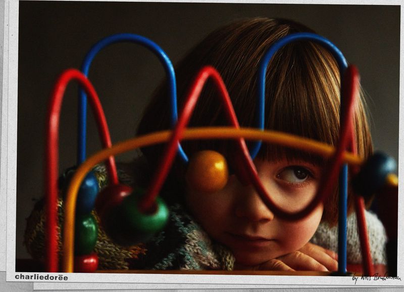 Close-up portrait of a child with short brown hair, resting their head on their hands, partially obscured by a colorful bead maze toy.