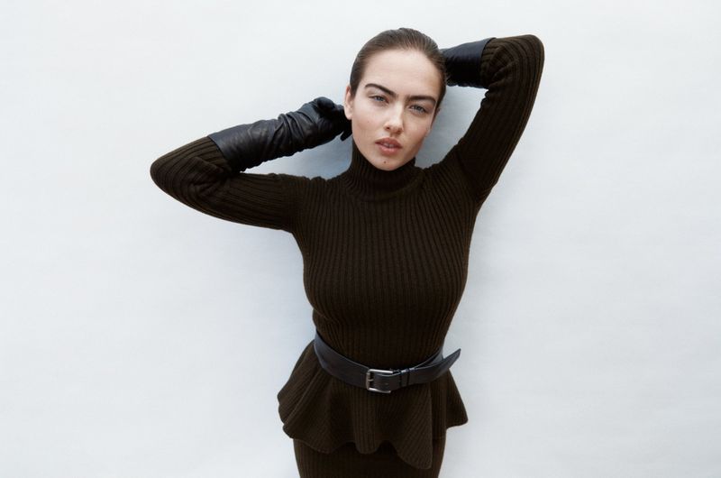 A fashionable woman in a dark green, ribbed turtleneck dress with leather gloves poses against a bright white wall in a studio setting.