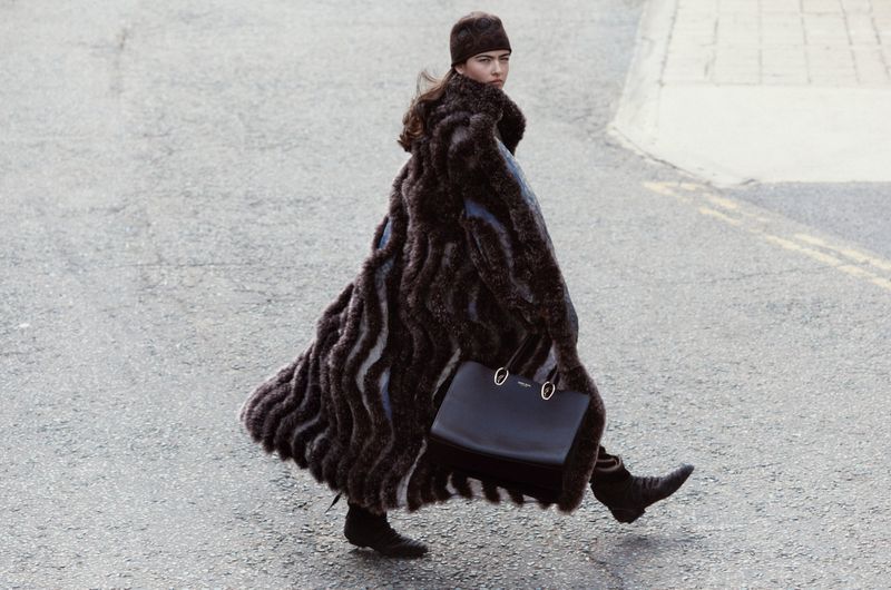 Woman in a long fur coat and matching hat crossing a street with a black tote bag, showcasing fashion and urban style in an outdoor setting.
