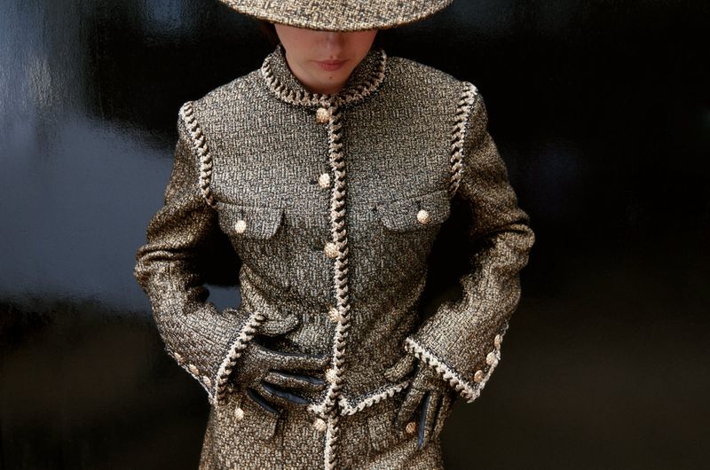 Woman in tweed jacket and hat against dark background. Textured fabric fashion shot with neutral palette, eye-level view, formalwear.