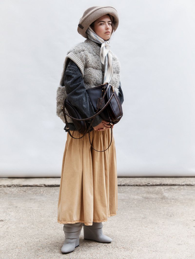 Full-shot of a woman wearing a hat, scarf, and skirt, with a large black purse, standing in front of a concrete and white backdrop.