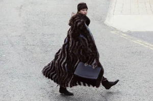 Model in textured brown coat and beanie walking on a paved street carrying a dark structured handbag
