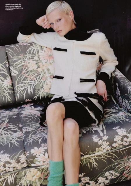 Attractive adult woman with platinum blonde hair in a black and white suit sitting on a floral couch, looking at the camera.