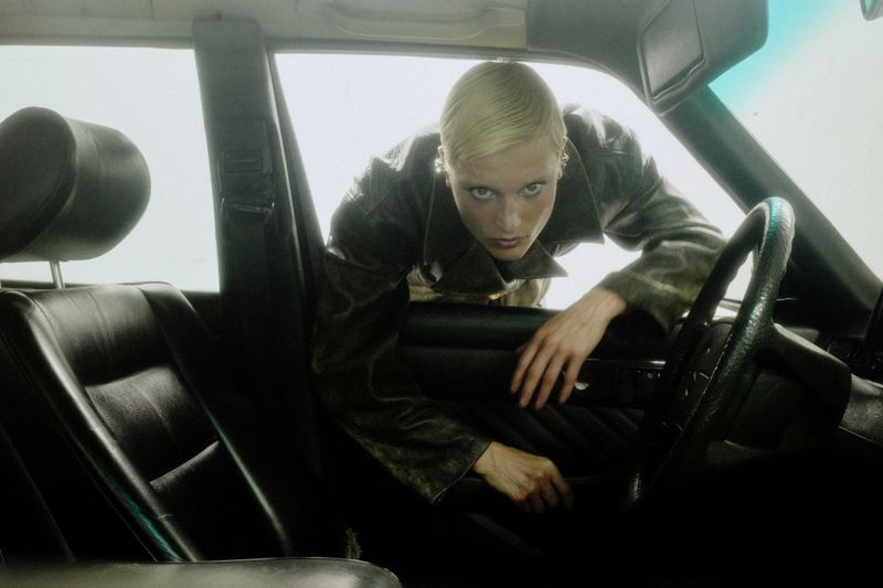 Blonde woman in vintage car leans out driver's side window, gazing intently with a neutral expression in natural light.