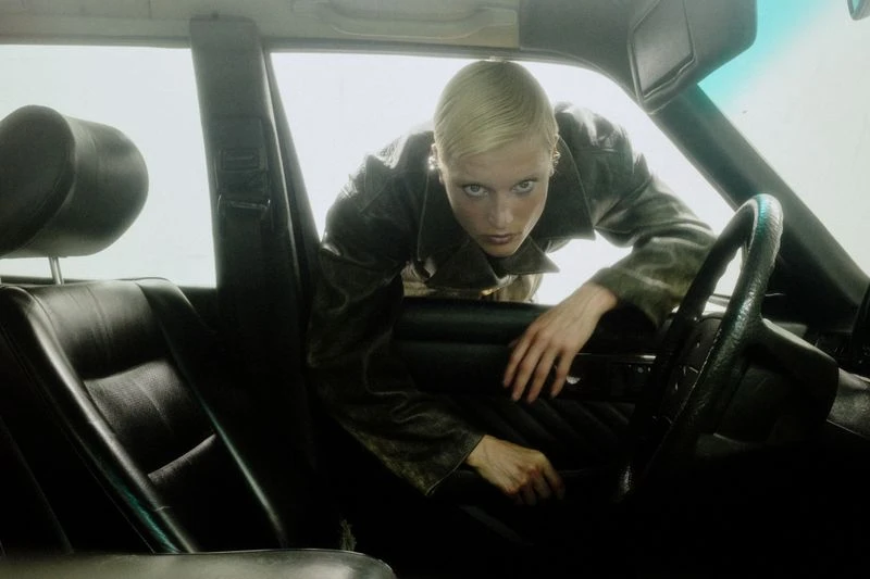 Blonde woman in vintage car leans out driver's side window, gazing intently with a neutral expression in natural light.