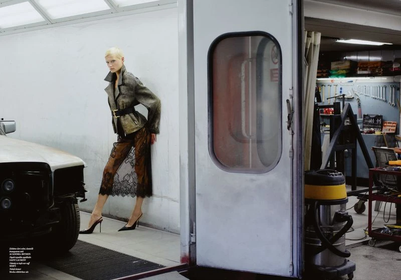 Fashion shot featuring a woman in a jacket and lace skirt, leaning against a wall in an auto body shop, with tools and car parts.