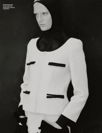 Monochromatic fashion editorial shot of a woman wearing a white boucle jacket with black accents against a dark background in a studio.