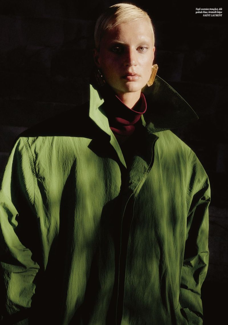 A blond woman with a green coat and golden earrings poses in soft lighting against a dark background for a fashion or beauty editorial.