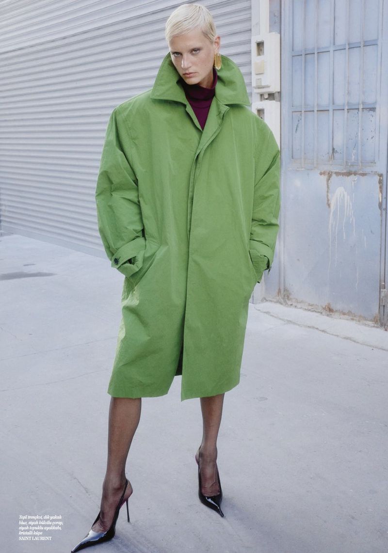Fashion editorial image of a blonde woman in a bright green oversized trench coat, black heels, and black tights against a gray wall.