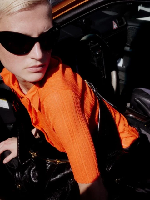 A woman with sunglasses leans out of a car. She has short blonde hair and wears an orange top. A black bag sits beside her.