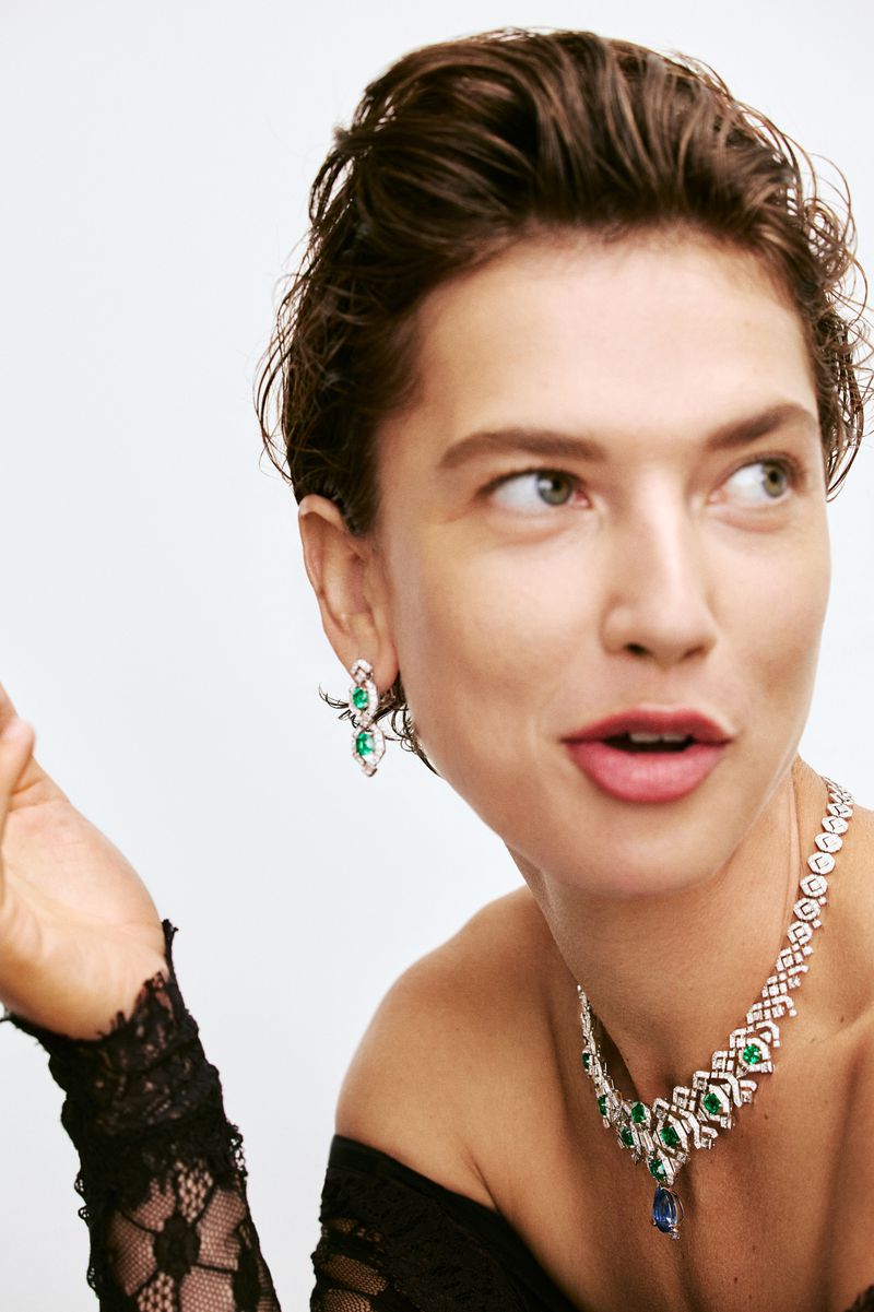 Elegant adult woman with short brunette hair wearing luxury diamond and green gemstone jewelry, black lace top, white background.