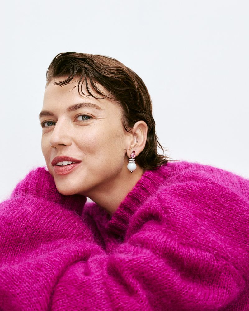 Close-up shot of an adult woman with short, wavy brown hair wearing a pink fuzzy sweater and pearl earrings in a white studio.