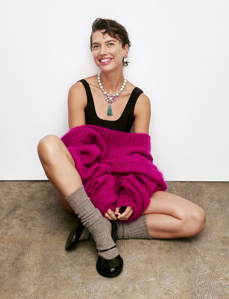 Model Gigi Ringel sitting cross-legged wearing a magenta knit sweater, black tank top, statement pearl necklace, and grey socks.