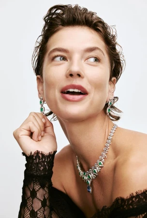 Adult female poses in a close-up wearing diamond and emerald earrings and necklace in a studio shot against a white backdrop.