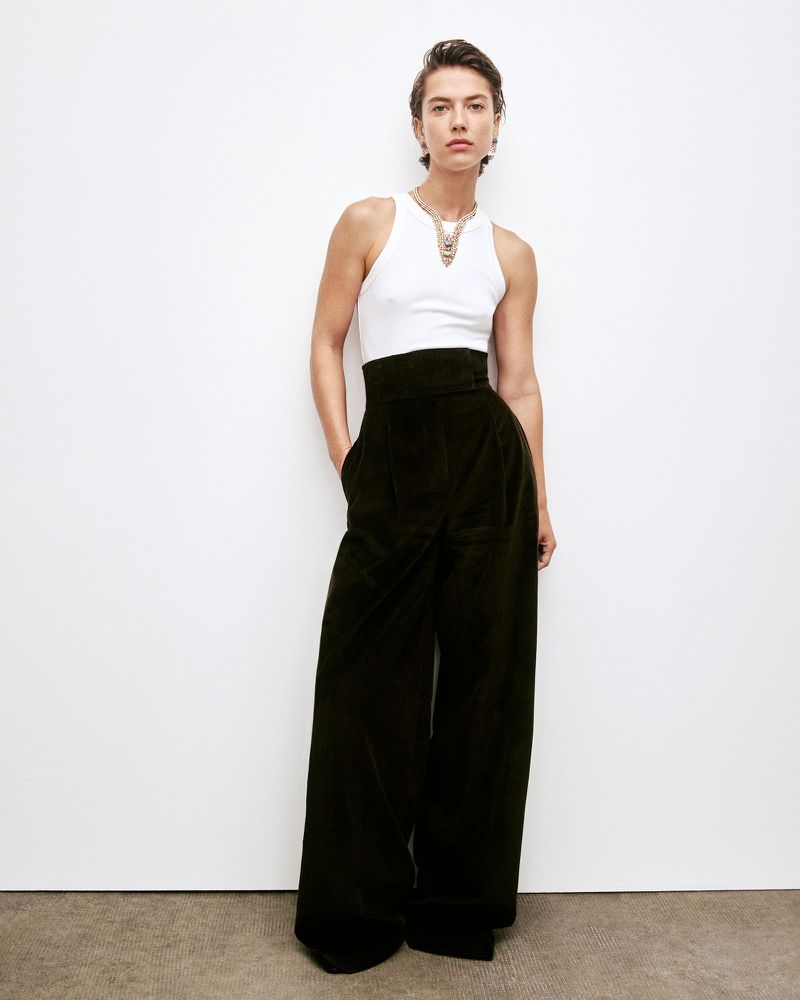 Fashion portrait of a woman wearing a white tank top, black wide-leg pants, and a gold necklace standing in front of a white wall.