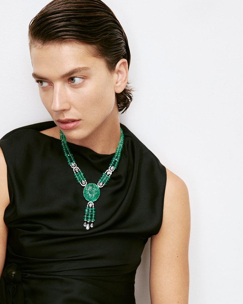 Close-up of a woman wearing a black dress and an elaborate green beaded necklace with a medallion and diamond accents.