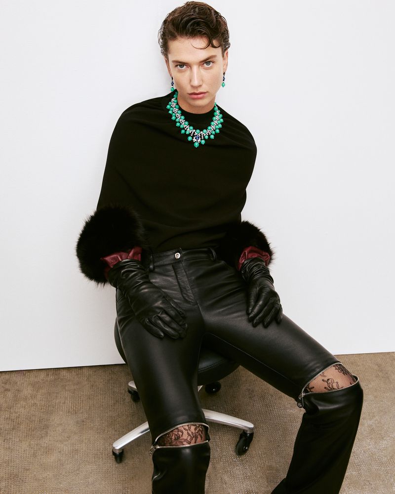 Woman in black top, leather pants, and boots poses with green jewelry against a white background in a fashion studio shot.