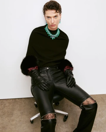 Woman in black top, leather pants, and boots poses with green jewelry against a white background in a fashion studio shot.