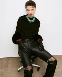 Model seated wearing a black top, leather trousers with knee zippers, leather gloves, and a bright green gemstone necklace.