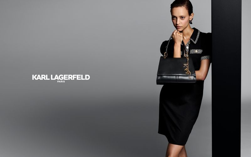Model in a black dress holding a black Karl Lagerfeld leather handbag with a gold charm against a grey studio background.