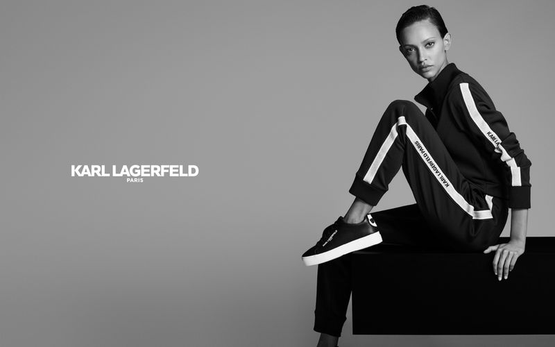 Fashion shot of a model wearing a Karl Lagerfeld branded tracksuit and sneakers, sitting against a gray background in a photo studio.