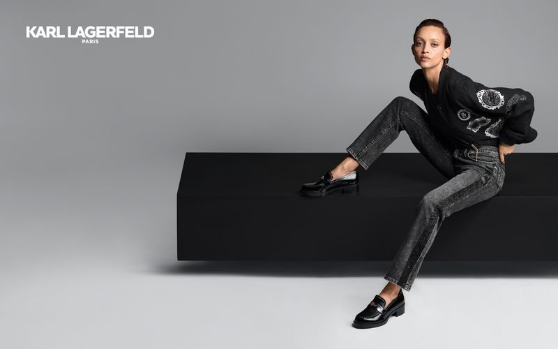Fashion editorial shot of a slim adult woman posing seated on a black block in gray jeans, black shoes, and a black Karl Lagerfeld sweater.