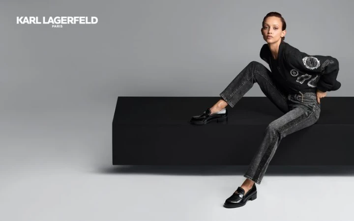 Fashion editorial shot of a slim adult woman posing seated on a black block in gray jeans, black shoes, and a black Karl Lagerfeld sweater.
