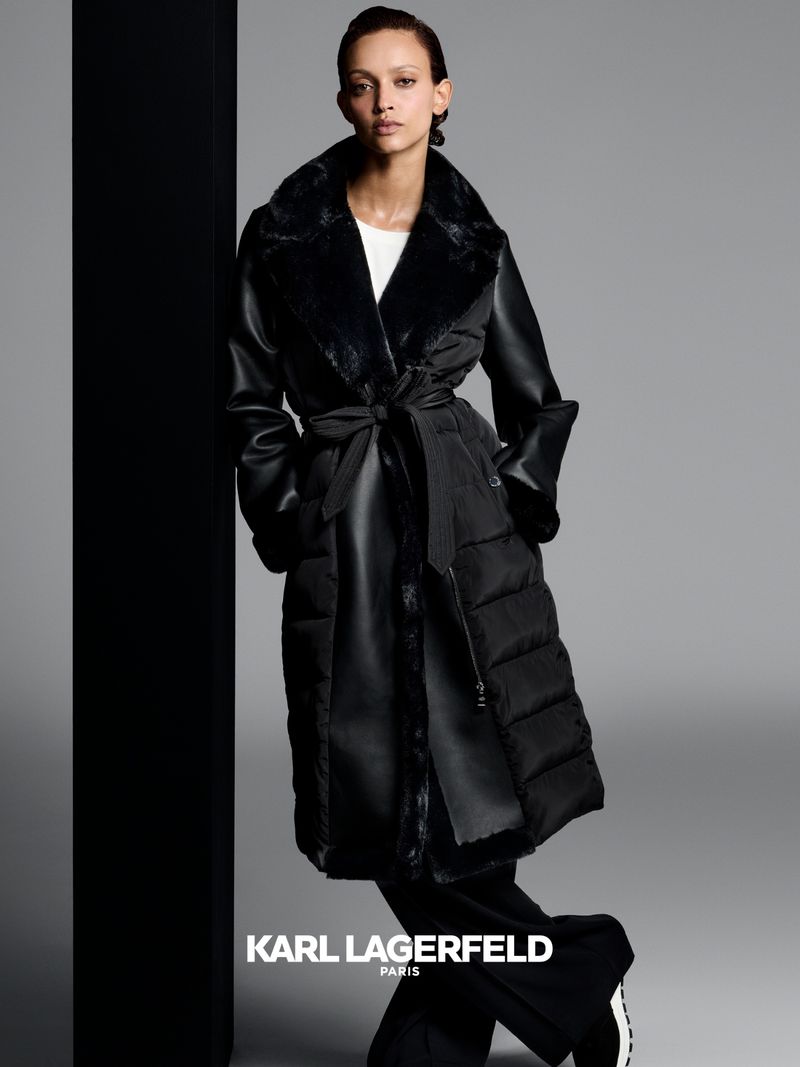 Woman poses in a black Karl Lagerfeld puffer coat with faux fur collar and wide leg pants against a grey and black background.