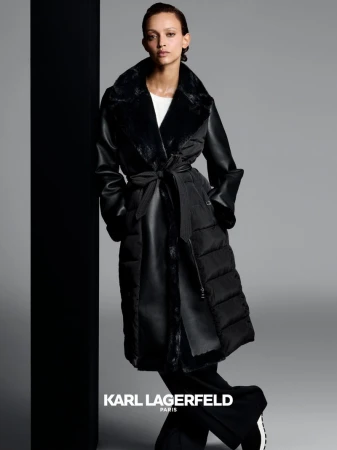 Woman poses in a black Karl Lagerfeld puffer coat with faux fur collar and wide leg pants against a grey and black background.