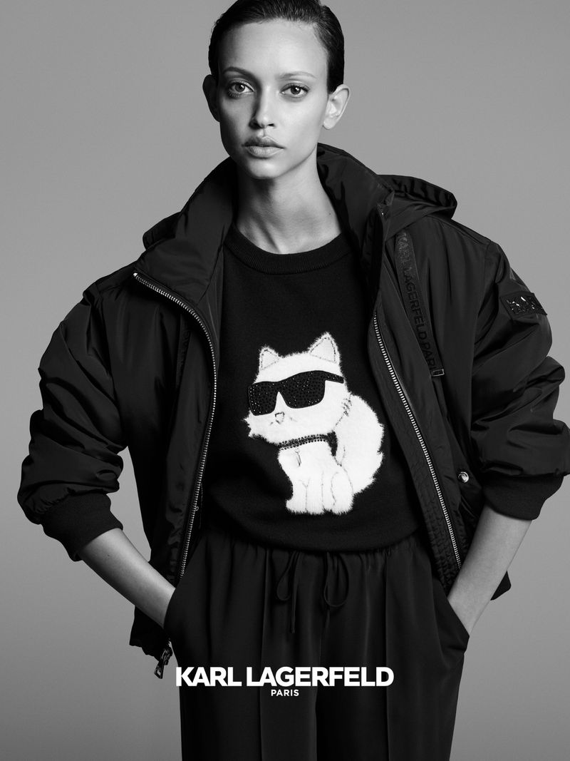 Fashion shot of a woman in a Karl Lagerfeld jacket and pants, posing in front of a gray background. The jacket features a hooded design.