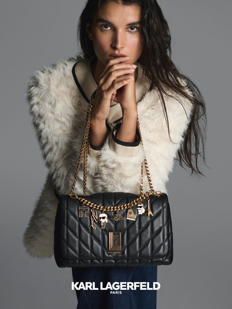 Woman poses with black Karl Lagerfeld Paris shoulder bag with gold chain and charms, wearing a white fur coat on gray background.