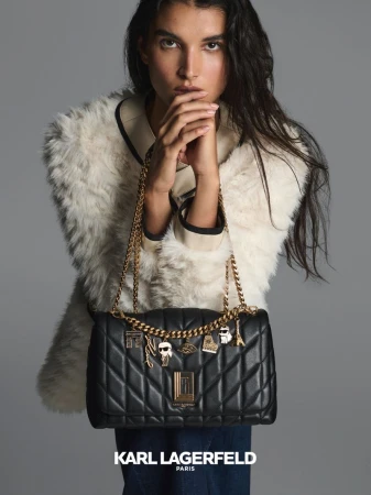 Woman poses with black Karl Lagerfeld Paris shoulder bag with gold chain and charms, wearing a white fur coat on gray background.