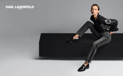 Model seated on a black block wearing a black jacket, dark jeans, and loafers against a grey seamless background