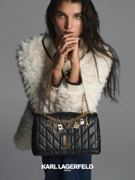 Model holding a black quilted leather handbag with gold chain charms against a neutral grey background.