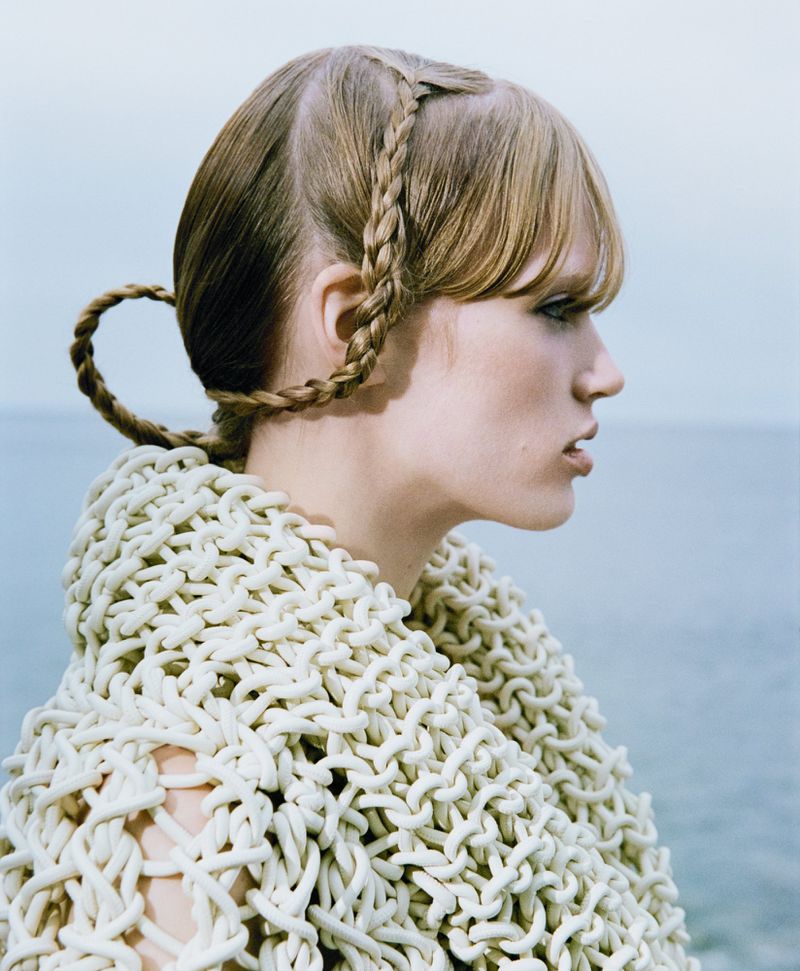 Woman with braided hair, wearing white knitted shawl stands against light blue sky and sea in outdoor fashion shot with shallow depth of field.