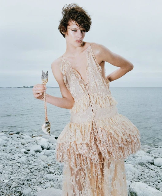 A person in a lace dress stands on a rocky beach near the ocean, holding a fish skeleton, under an overcast sky in a fashion editorial.