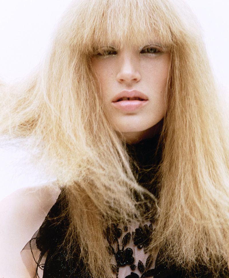 Close-up portrait of a blonde model with heavy textured bangs wearing a black embellished top against a bright white background.