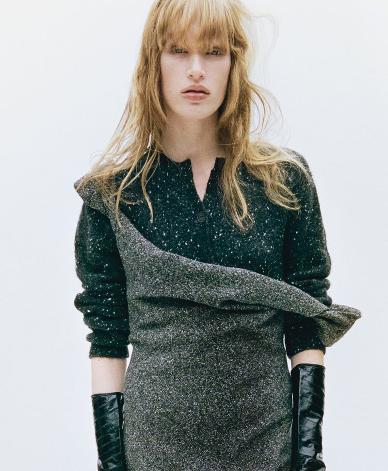 An adult woman in a dress poses indoors, showcasing a stylish ensemble in a close-up shot with a light background.