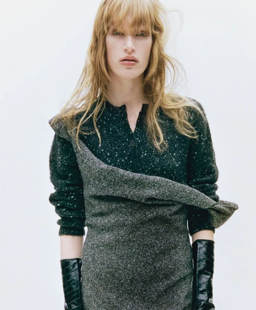 An adult woman in a dress poses indoors, showcasing a stylish ensemble in a close-up shot with a light background.