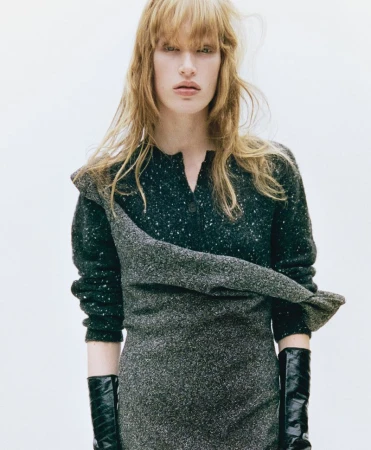 An adult woman in a dress poses indoors, showcasing a stylish ensemble in a close-up shot with a light background.