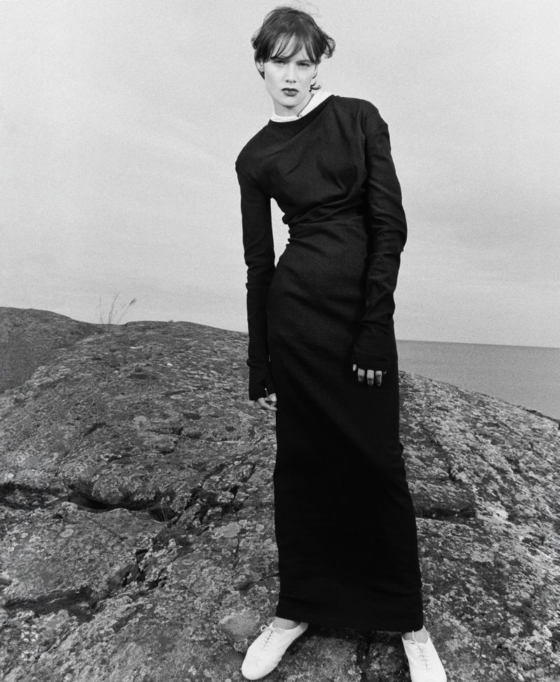 Model wearing a long black long-sleeved dress with white shoes while posing on a rocky coastal ridge in a black and white editorial.