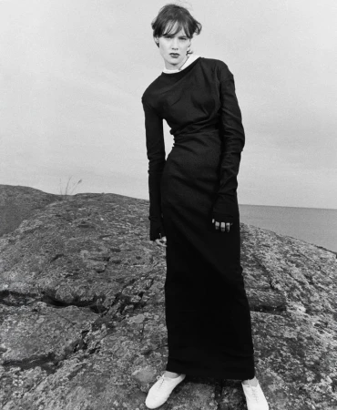 Model wearing a long black long-sleeved dress with white shoes while posing on a rocky coastal ridge in a black and white editorial.