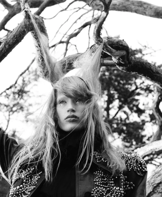 Monochrome portrait of a model with glitter makeup, wearing a studded jacket, posed amongst tree branches in a low angle shot.