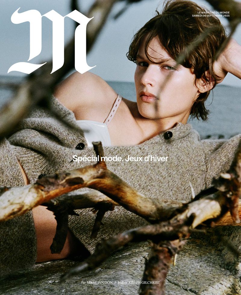 Fashion magazine cover with a woman in a neutral-toned sweater lying amongst driftwood against a grey sky backdrop.