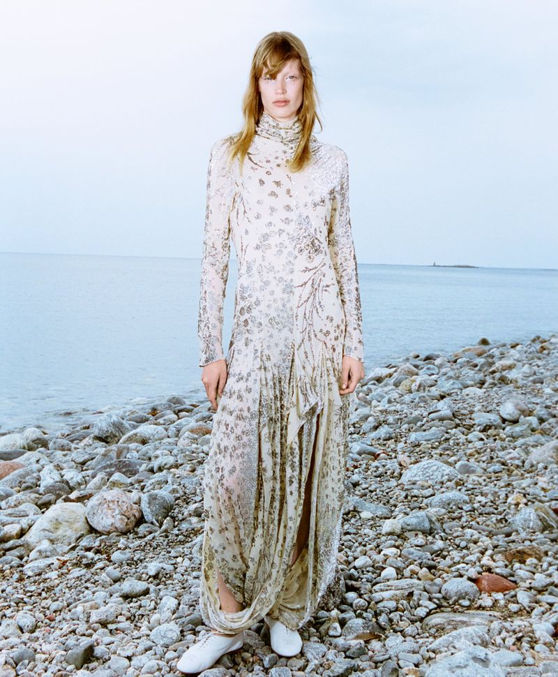 Woman in flowing floral gown on a rocky beach by the ocean. Full-length shot with pale blue water and sky background.